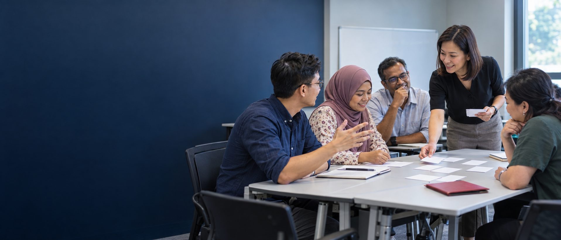 A trainer smiling as she hands out cards to five adult learners at a table in a classroom, one of them laughing and explaining something with his hands.
