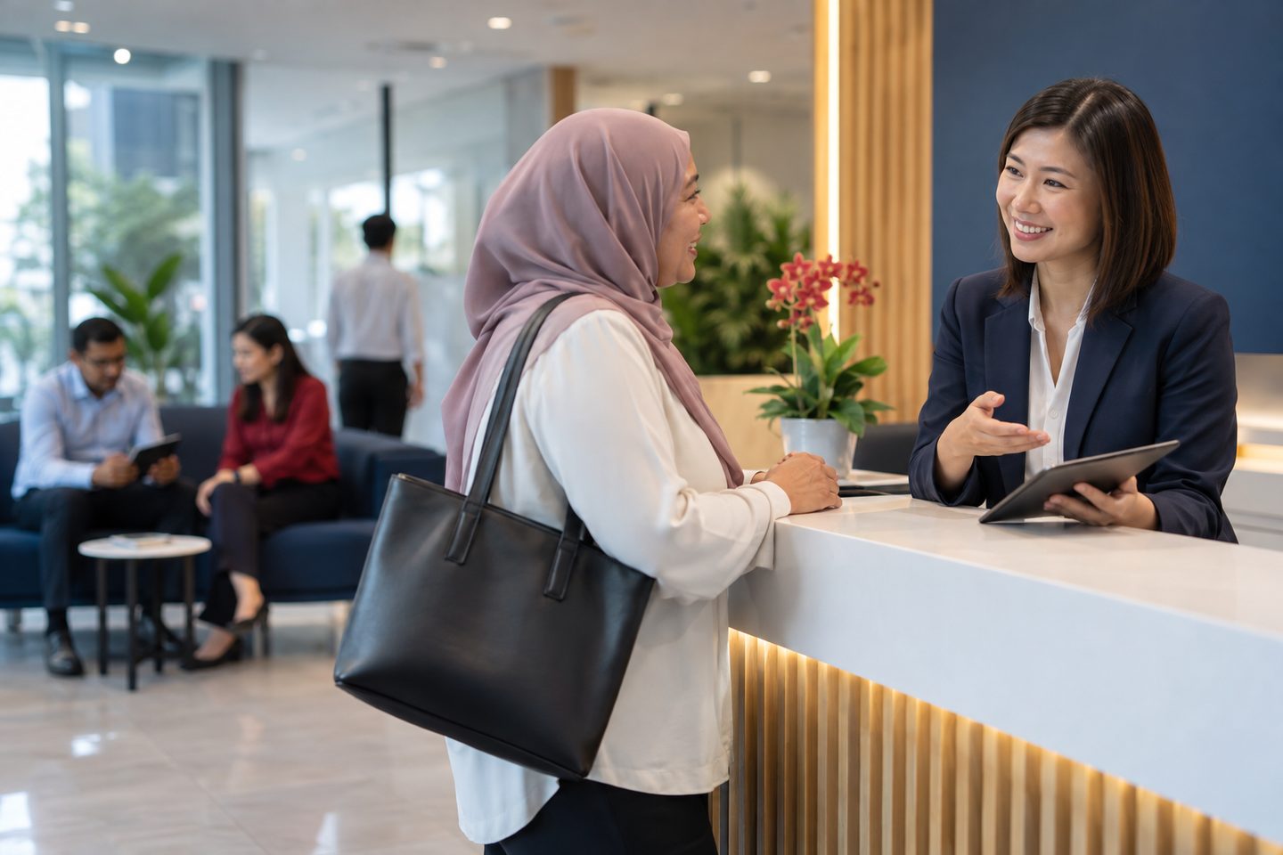 A woman in a pink headscarf talking with a smiling receptionist in a navy blazer, who holds a tablet across a white reception counter, people seated in a lounge behind them.