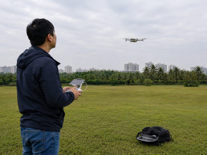 A person flying a drone with a handheld controller over an open field, the drone hovering ahead of them.